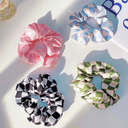 Flat lay of four satin checkerboard scrunchies in pink, blue, black, and green.