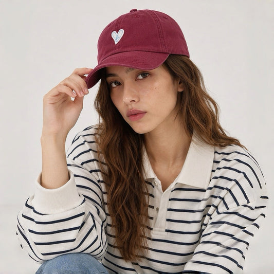 Female model wearing a burgundy cotton baseball cap with white heart embroidery paired with a striped polo.