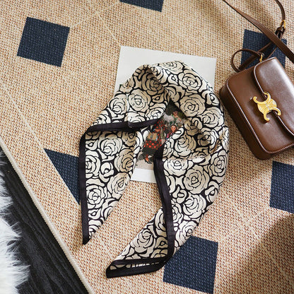 Angled view of Rose silk scarf, with black and white rose pattern, on a patterned rug, easy for travel.