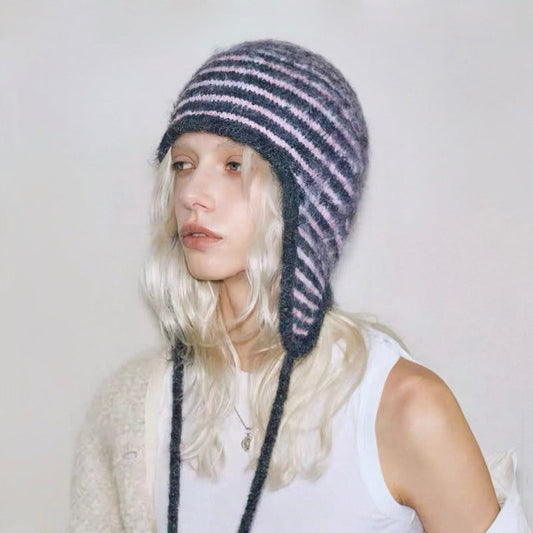 Model wearing a pink and dark grey striped knit earflap hat with braided tassel ties.